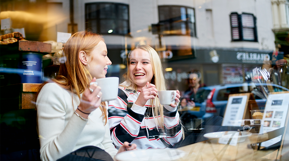 Two teenage female friends enjoying a coffee and a moment together sitting in a cafe window 